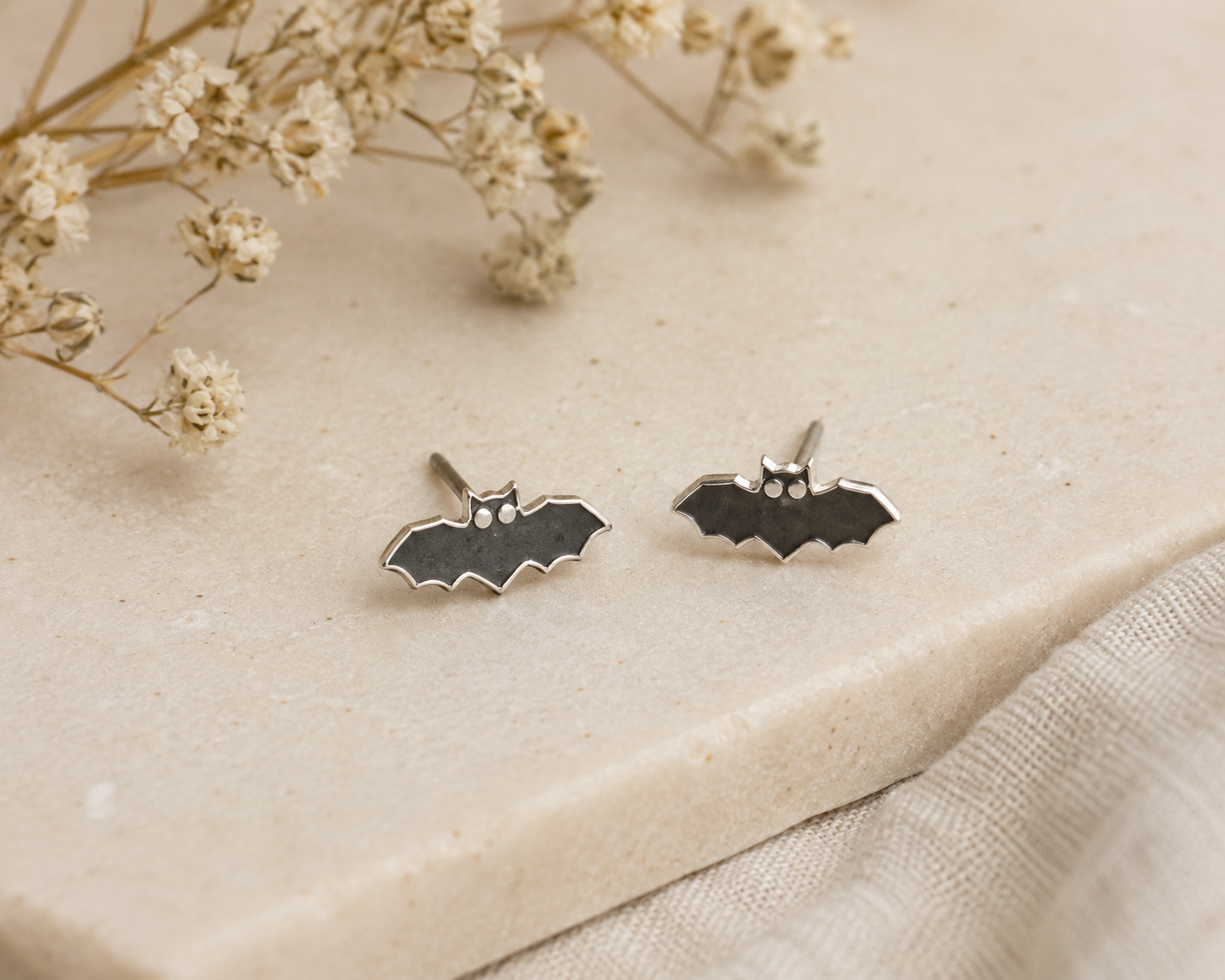 Bat Stud Earrings Sterling Silver — pair of small black enamel bat shaped stud earrings with silver outline on cream stone surface with dried gypsophila"