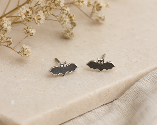 Bat Stud Earrings Sterling Silver — pair of small black enamel bat shaped stud earrings with silver outline on cream stone surface with dried gypsophila"