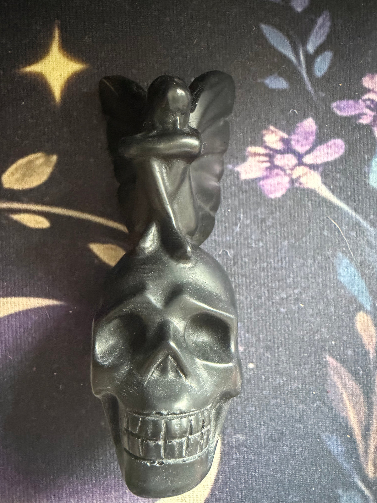 A black gothic figurine featuring a winged angel sitting atop a detailed skull, displayed on a dark patterned fabric background.