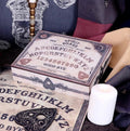 A vintage-style wooden spirit board and card deck set displayed on a dark surface, including a square spirit board box, a black heart-shaped card, and a white pillar candle.