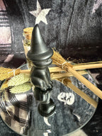 A small matte black witch figurine with a pointed hat, displayed on a glass plate with a broomstick and dried grasses against a dark plaid fabric background.