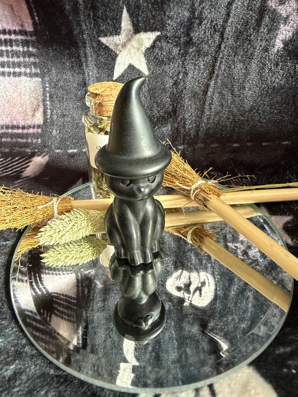 A small matte black witch figurine with a pointed hat stands on a round mirrored tray, styled with dried broomsticks, wheat, and a small glass bottle against a dark plaid fabric background.