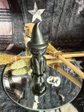 A small matte black witch figurine with a pointed hat stands on a round mirrored tray, styled with dried broomsticks, wheat, and a small glass bottle against a dark plaid fabric background.