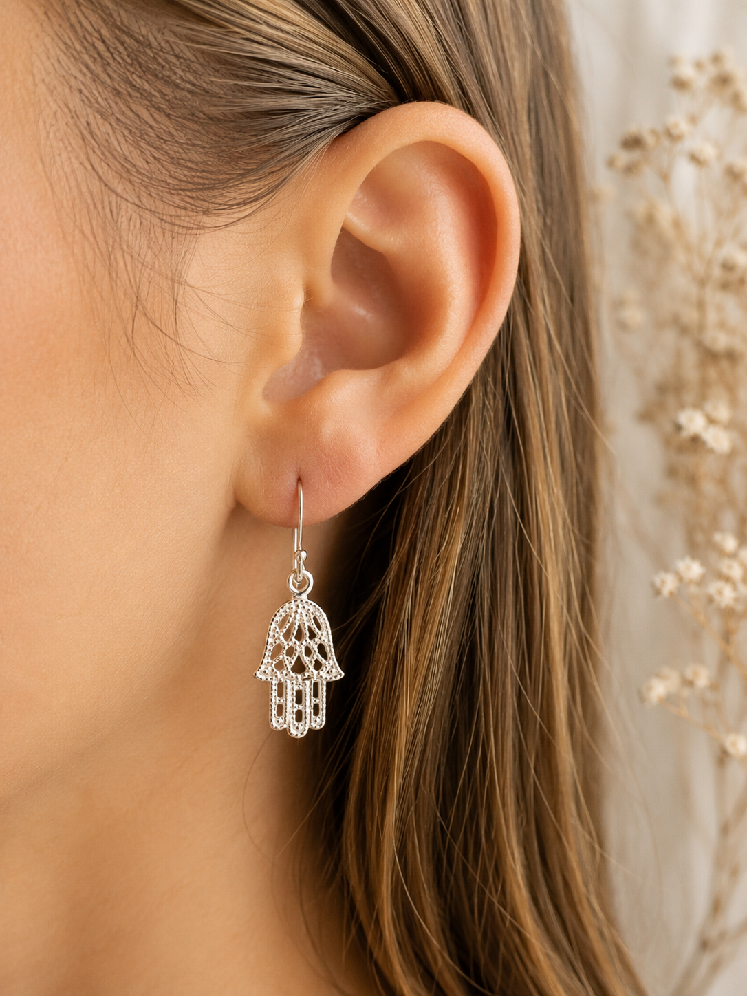 "Hamsa Hand Drop Earrings Sterling Silver worn shot — single silver hook earring with delicate filigree openwork hamsa hand charm shown worn on model's ear with dried gypsophila in background"