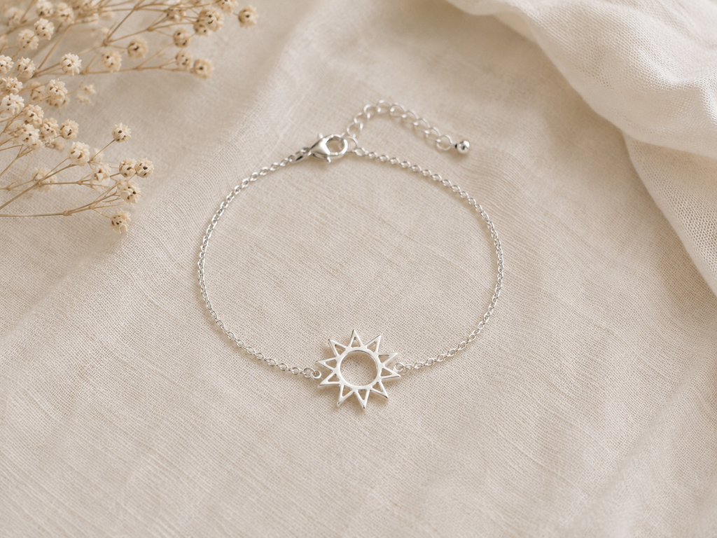 A delicate silver chain bracelet with a sun-shaped charm, displayed on a textured beige fabric background with dried baby's breath flowers in the corner.