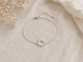 A delicate silver chain bracelet with a sun-shaped charm, displayed on a textured beige fabric background with dried baby's breath flowers in the corner.