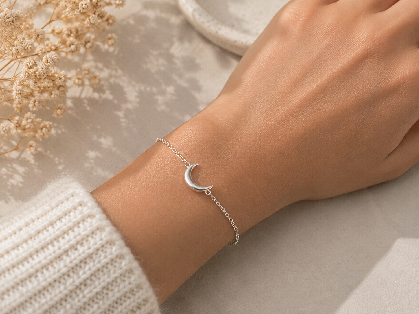 A delicate silver crescent moon chain bracelet worn on a person's wrist, styled with a white knit sweater sleeve and dried flowers on a neutral beige surface.