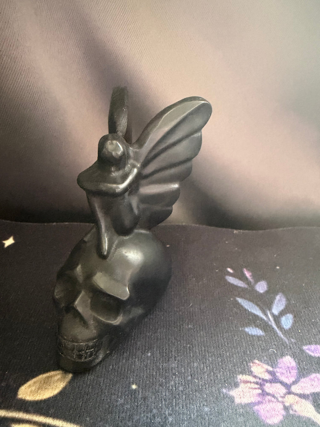 A matte black metal gothic winged skull figurine, photographed on a dark patterned fabric surface against a dark gray background.
