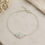 "Hamsa Hand Bracelet Sterling Silver product shot — delicate silver chain bracelet with pavé CZ hamsa hand charm and turquoise centre stone, spring ring clasp and extender chain, on cream marble surface with dried gypsophila"