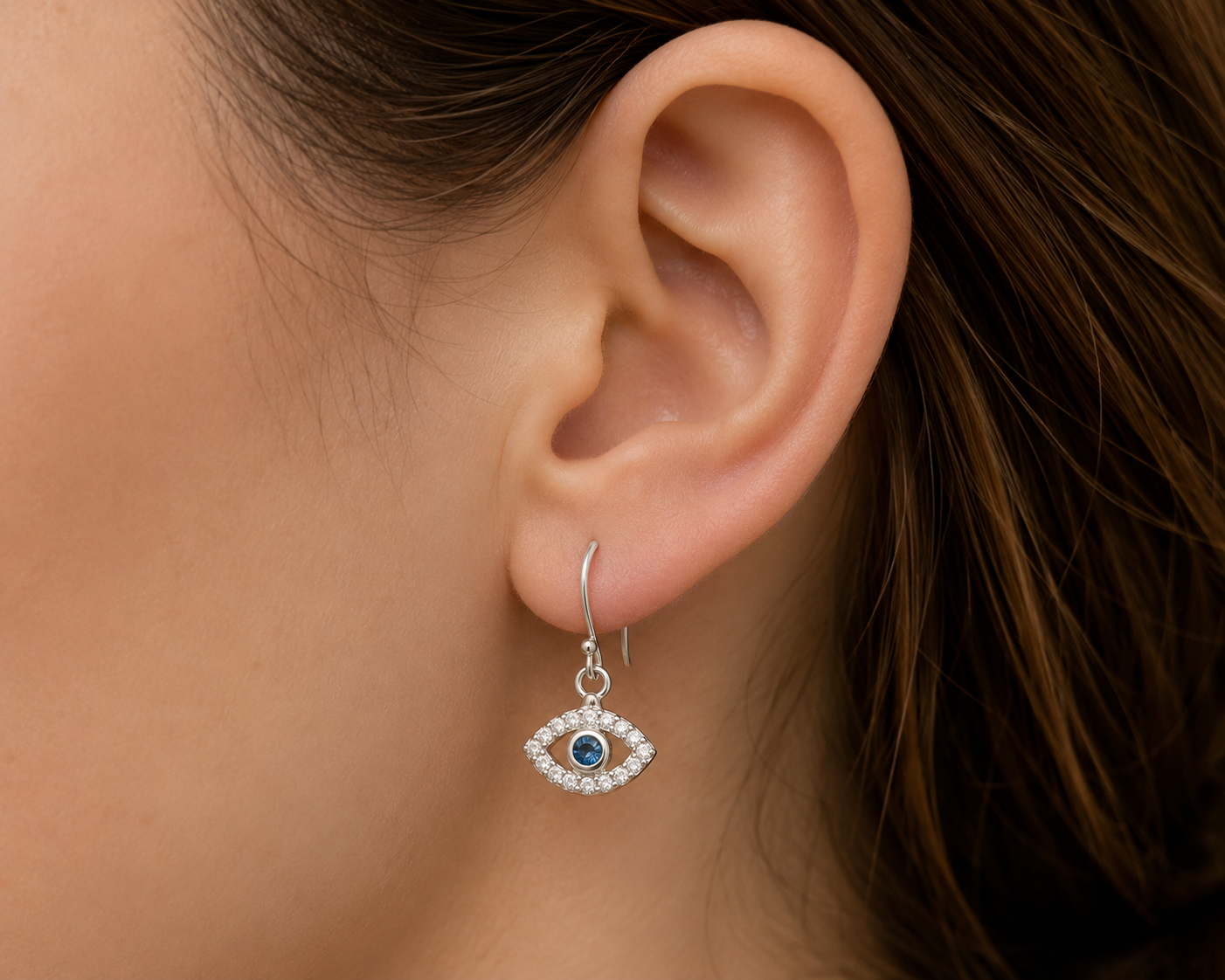 A close-up of a person's ear wearing a silver hook earring with an evil eye charm. The charm features a blue center surrounded by small clear stones.