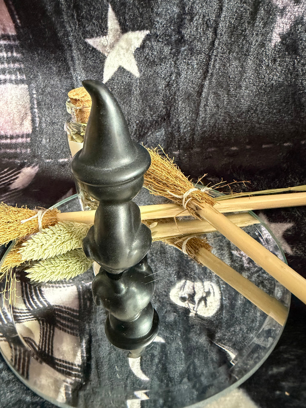 A black witch-style candle holder with a pointed hat finial, displayed on a glass tray with broomsticks and dried grass against a dark patterned fabric background.