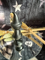 A black witch-style candle holder with a pointed hat finial, displayed on a glass tray with broomsticks and dried grass against a dark patterned fabric background.