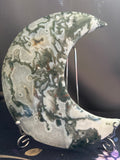 A crescent moon-shaped decorative stone plaque with a marbled agate pattern in shades of green, gray, white, and brown, displayed on a small silver stand.