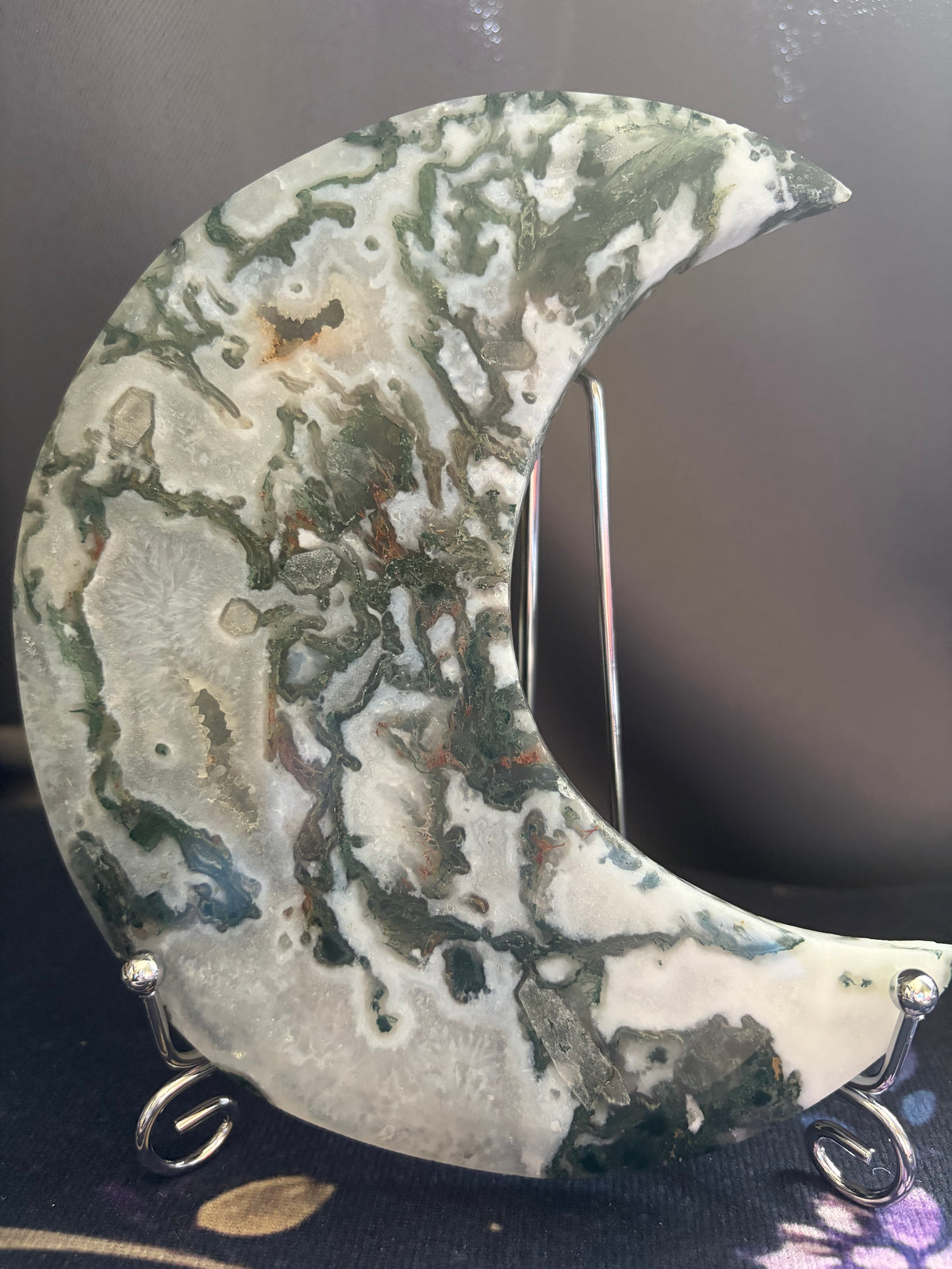 A crescent moon-shaped decorative stone plaque with a marbled agate pattern in shades of green, gray, white, and brown, displayed on a small silver stand.