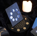 A tarot card set displayed on a black crescent moon stand featuring a gold flower of life design and moon phases. The main card shows a full moon and moon phase sequence, set against a dark velvet background with a glowing salt lamp in the background.