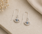 A pair of silver-tone drop earrings with an eye design, featuring a central blue gemstone surrounded by small clear crystals, displayed on a beige stone surface with dried flowers and a beige fabric in the background.