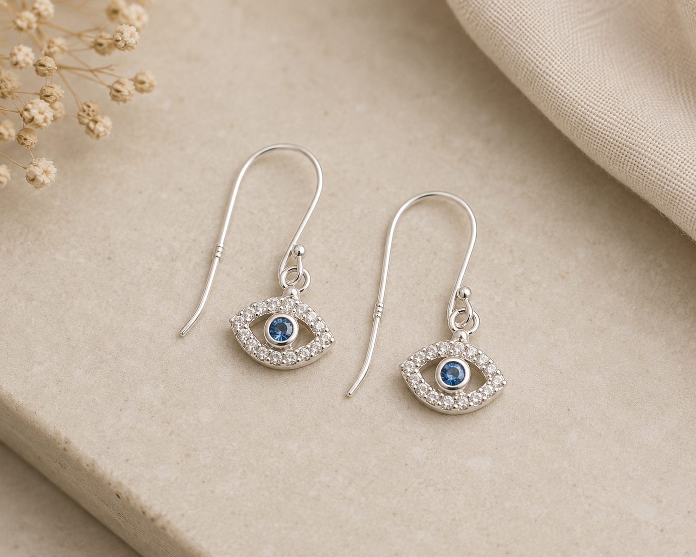 A pair of silver-tone drop earrings with an eye design, featuring a central blue gemstone surrounded by small clear crystals, displayed on a beige stone surface with dried flowers and a beige fabric in the background.