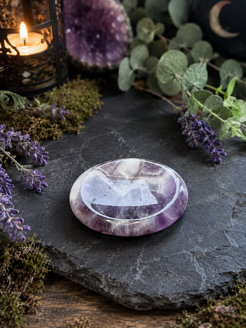 A polished round amethyst crystal plate with purple and white marbling, resting on a dark slate stone surrounded by green eucalyptus leaves, purple lavender sprigs, moss, and a lit candle in a lantern.