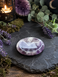 A polished round amethyst crystal plate with purple and white marbling, resting on a dark slate stone surrounded by green eucalyptus leaves, purple lavender sprigs, moss, and a lit candle in a lantern.