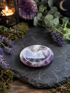 A polished round amethyst crystal plate with purple and white marbling, resting on a dark slate stone surrounded by green eucalyptus leaves, purple lavender sprigs, moss, and a lit candle in a lantern.