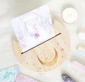 A wooden crescent moon card holder with laser-engraved crystal and star motifs, holding a card, styled on a soft white surface with crystals and a candle.