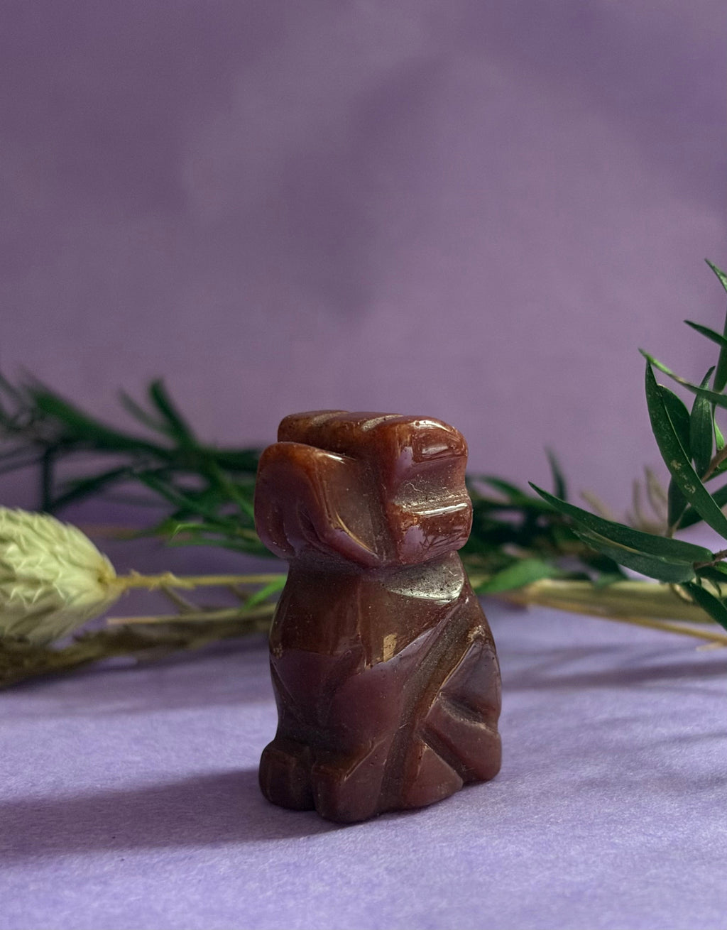 Red Goldstone crystal dog carving side view on lilac background with dried botanicals — The Crooked Moon