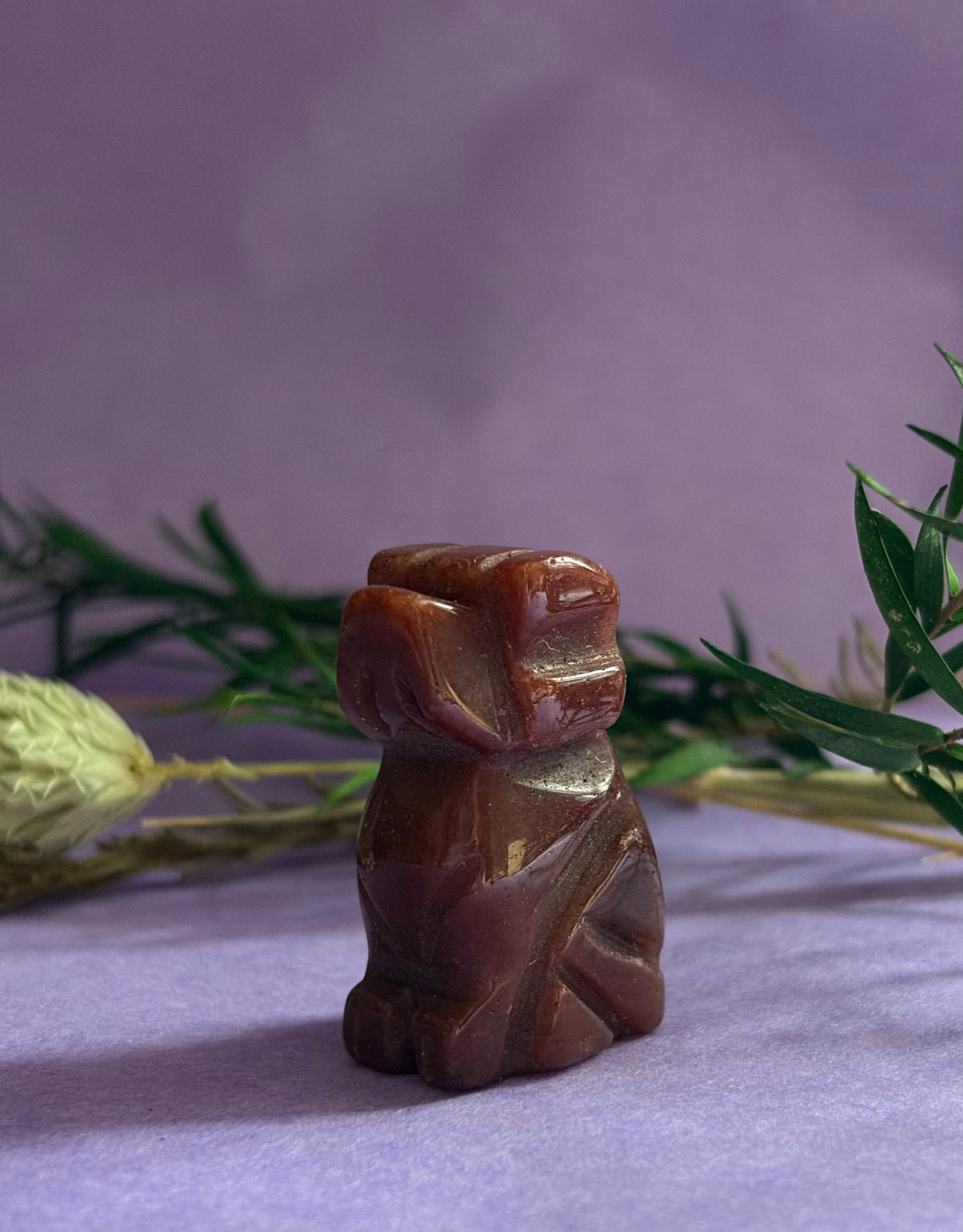 Red Goldstone crystal dog carving side view on lilac background with dried botanicals — The Crooked Moon