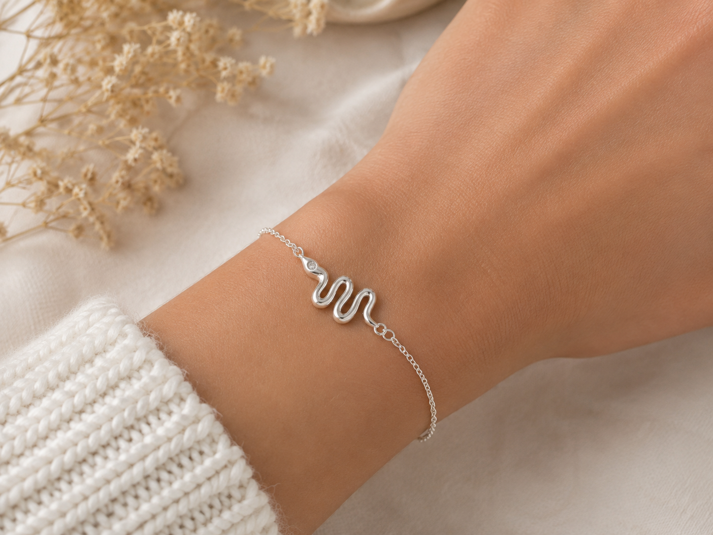 "Snake Chain Bracelet Sterling Silver worn shot — delicate silver chain bracelet with polished serpentine snake charm shown worn on model's wrist over white knit sleeve, dried gypsophila and white linen in background"