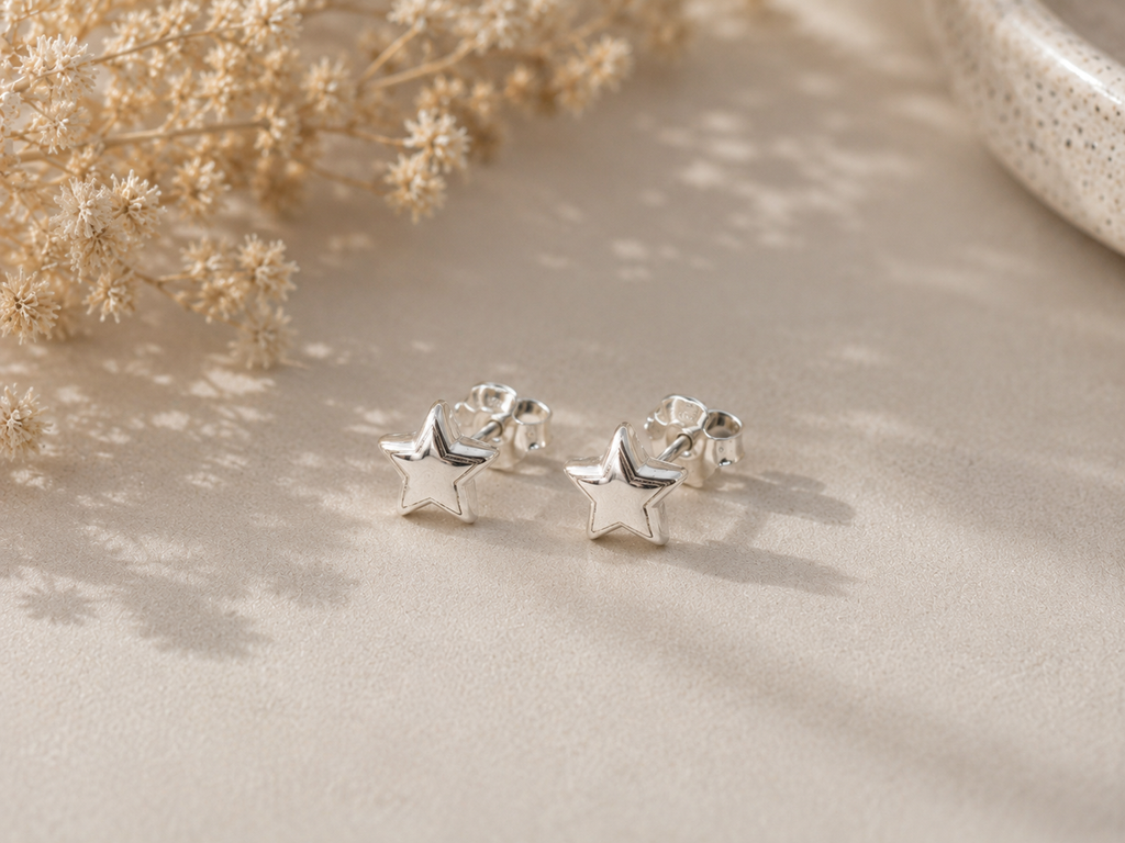 A pair of small silver star-shaped stud earrings resting on a beige textured surface with soft shadows, dried flowers, and a ceramic bowl in the background.