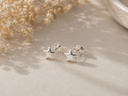 A pair of small silver star-shaped stud earrings resting on a beige textured surface with soft shadows, dried flowers, and a ceramic bowl in the background.