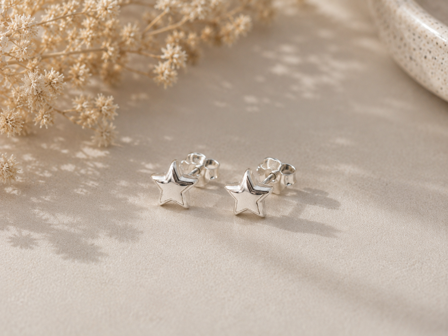 A pair of small silver star-shaped stud earrings resting on a beige textured surface with soft shadows, dried flowers, and a ceramic bowl in the background.