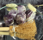 Five polished purple amethyst tumbled crystals arranged in a clear glass bowl, styled with dried wheat stalks and a small natural-fiber broom on wooden handles.