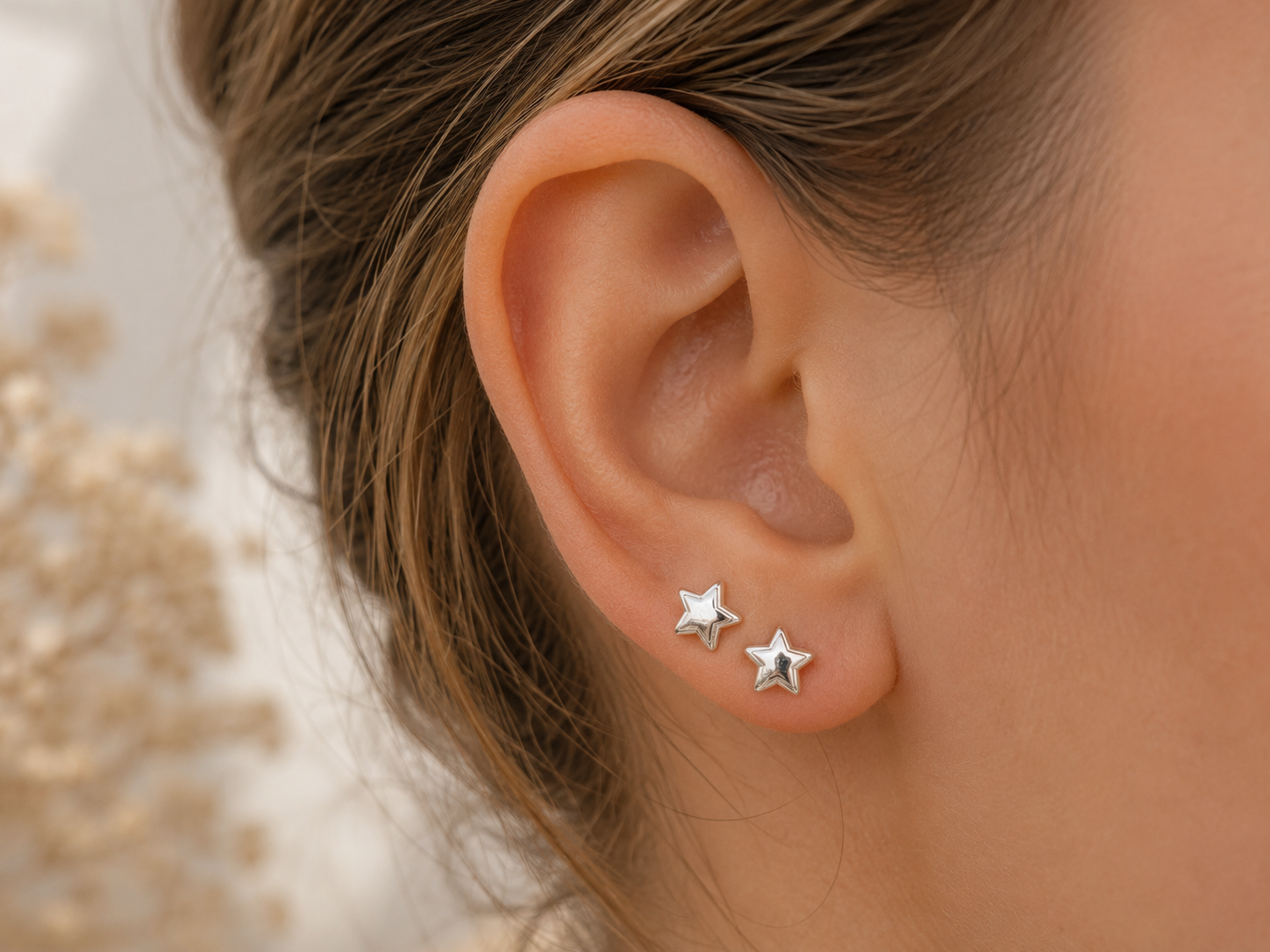 "Puffed Star Stud Earrings Sterling Silver worn shot — two polished silver puffed star studs of the same size shown worn in double-pierced ear lobe with dried gypsophila in background"