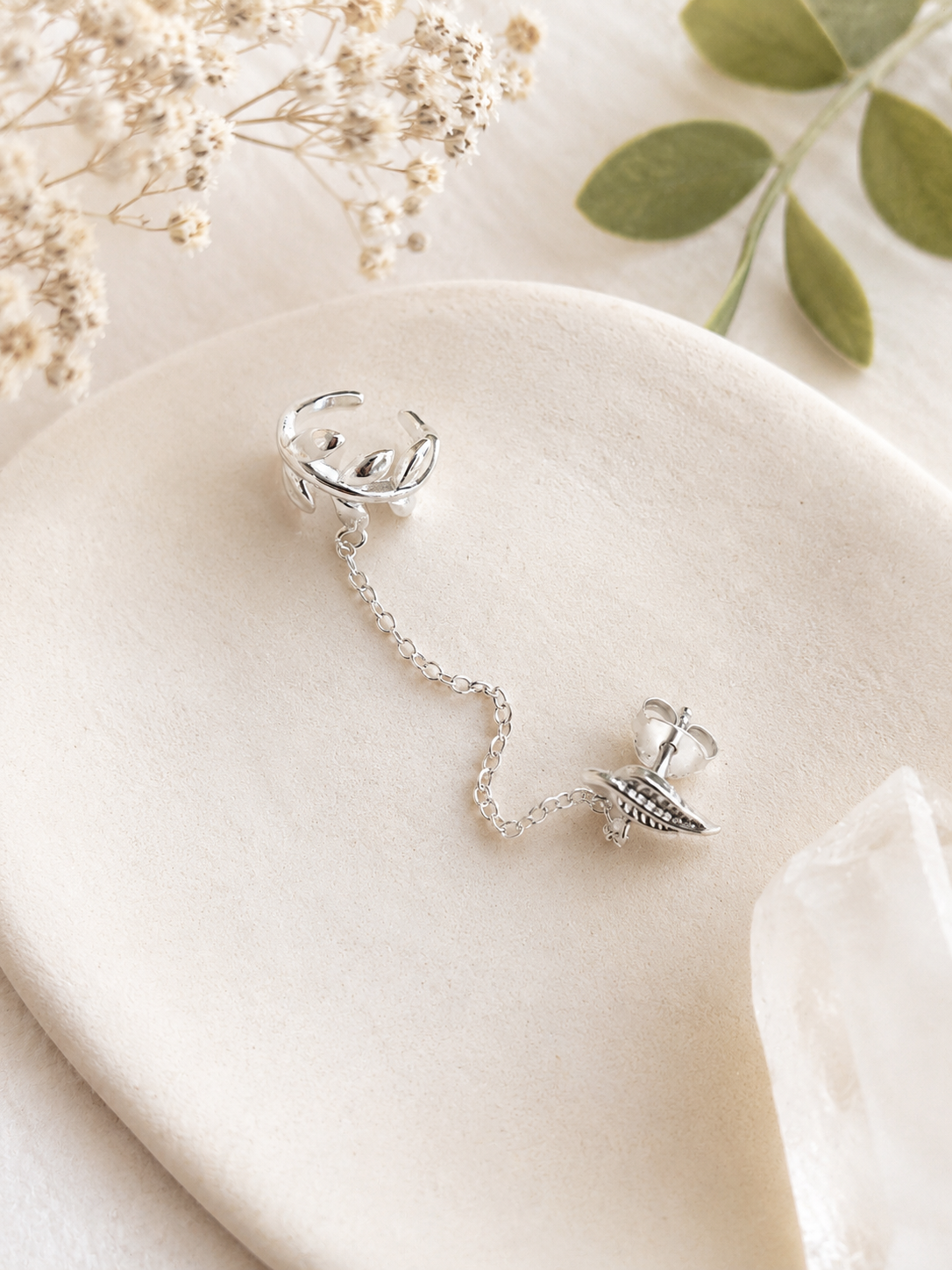 "Leaf Chain Ear Cuff & Stud Set Sterling Silver product shot — silver leaf ear cuff connected by a delicate chain to a small engraved leaf stud earring, displayed on cream ceramic dish with dried gypsophila and eucalyptus leaves"