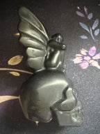 A black winged fairy figurine perched on top of a skull, photographed on a dark floral-patterned fabric background.