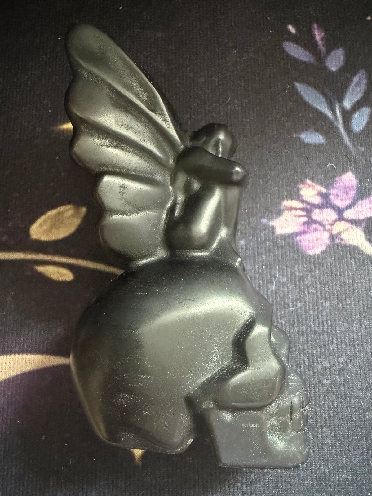 A black winged fairy figurine perched on top of a skull, photographed on a dark floral-patterned fabric background.