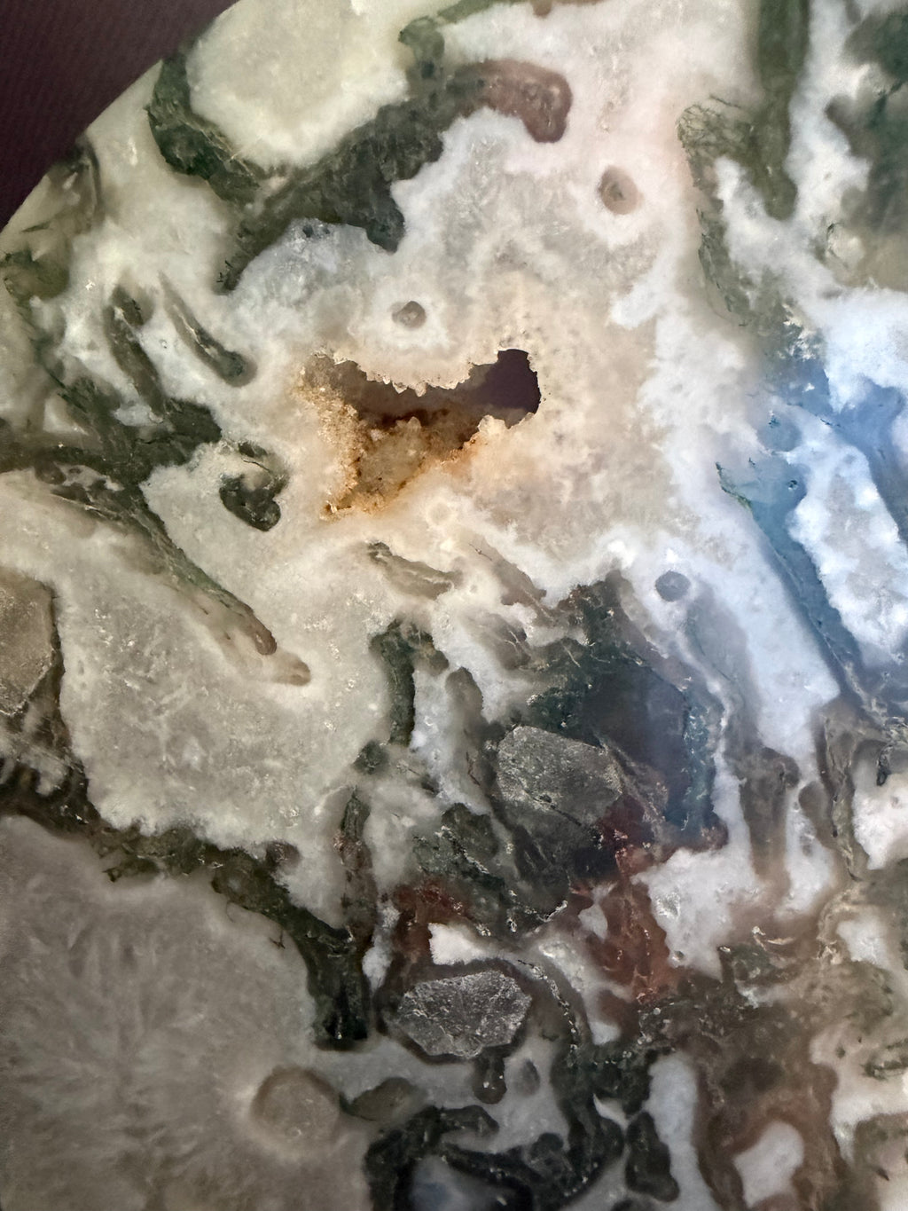 A close-up of a polished stone or mineral slab with marbled patterns in off-white, gray, olive green, and small brown areas, including a small dark cavity near the center.