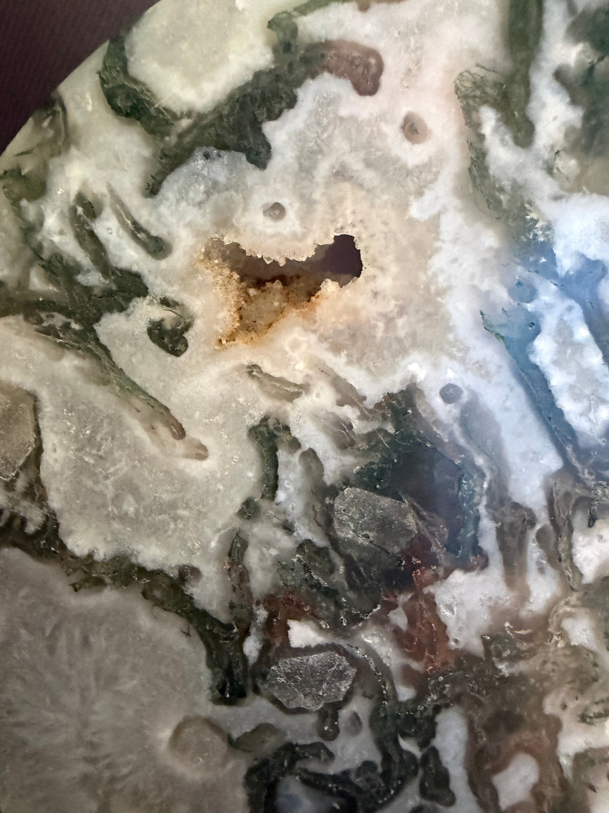 A close-up of a polished stone or mineral slab with marbled patterns in off-white, gray, olive green, and small brown areas, including a small dark cavity near the center.