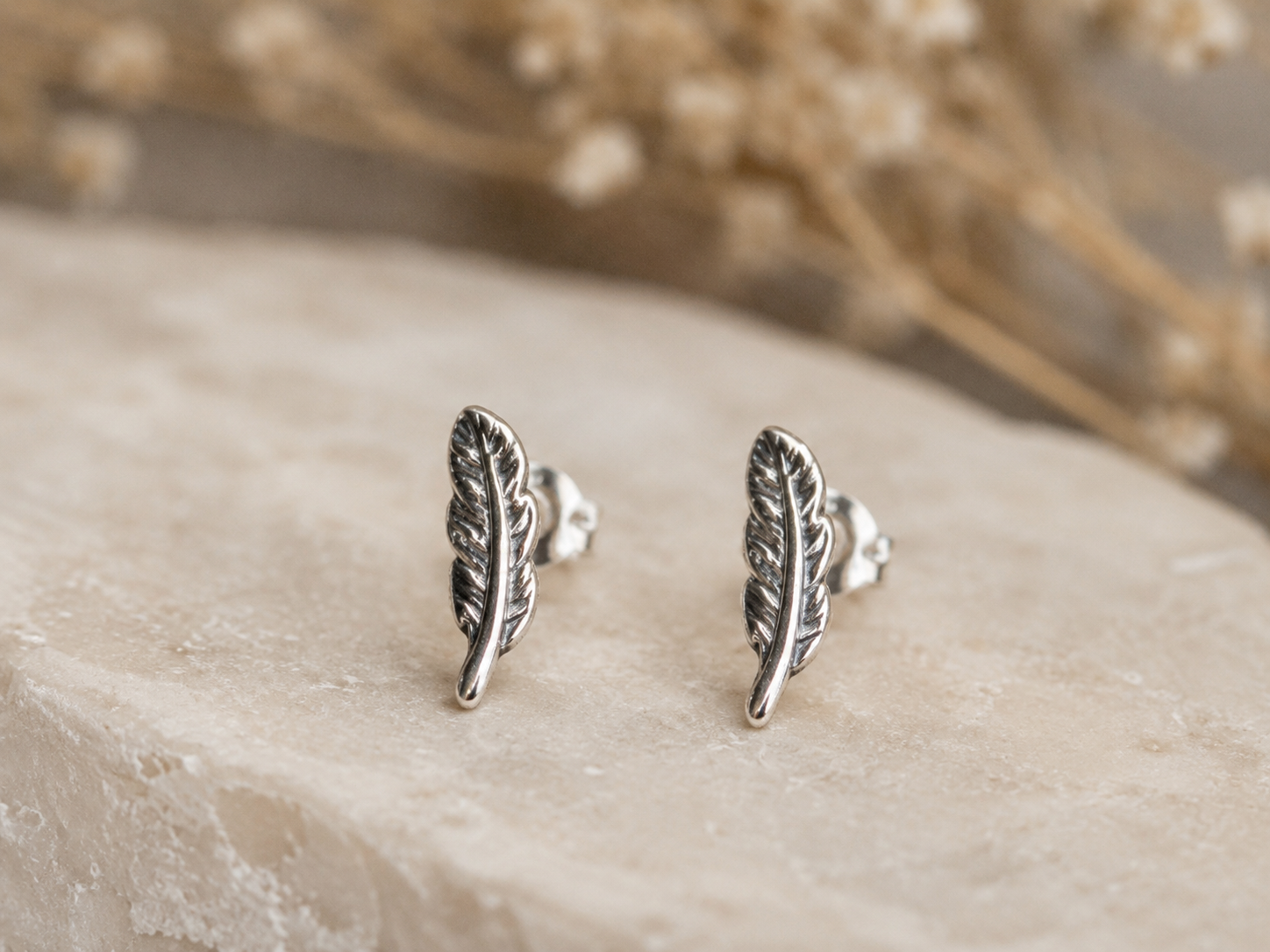A pair of silver feather stud earrings displayed on a light beige stone surface with a soft-focus background of dried flowers.
