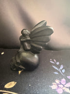 A small black figurine of a winged fairy or angel seated on a dark patterned fabric surface against a dark draped backdrop.