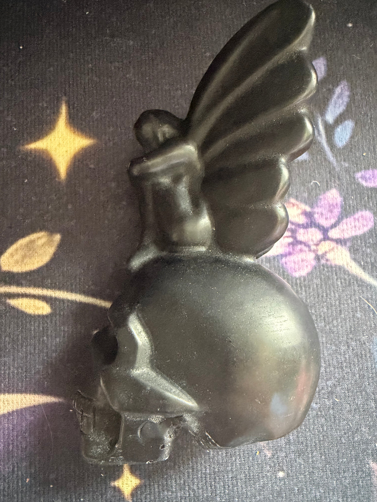 A black winged skull figurine or candle holder with a winged figure seated on top, photographed on a dark patterned fabric background.