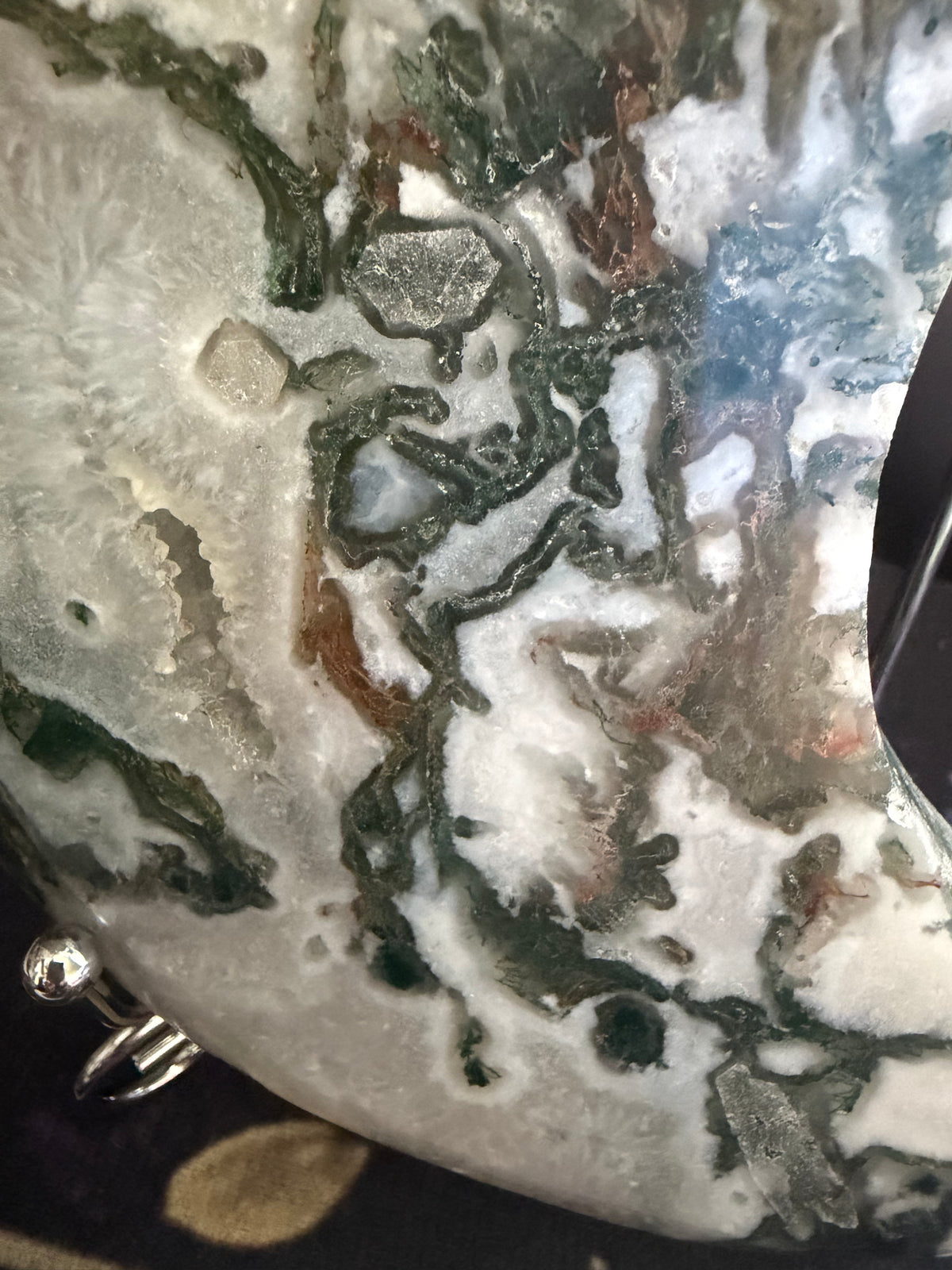 A close-up of a polished stone or agate slice with swirling patterns of white, gray, dark green, and brown, with a small silver metal setting visible at the bottom left.