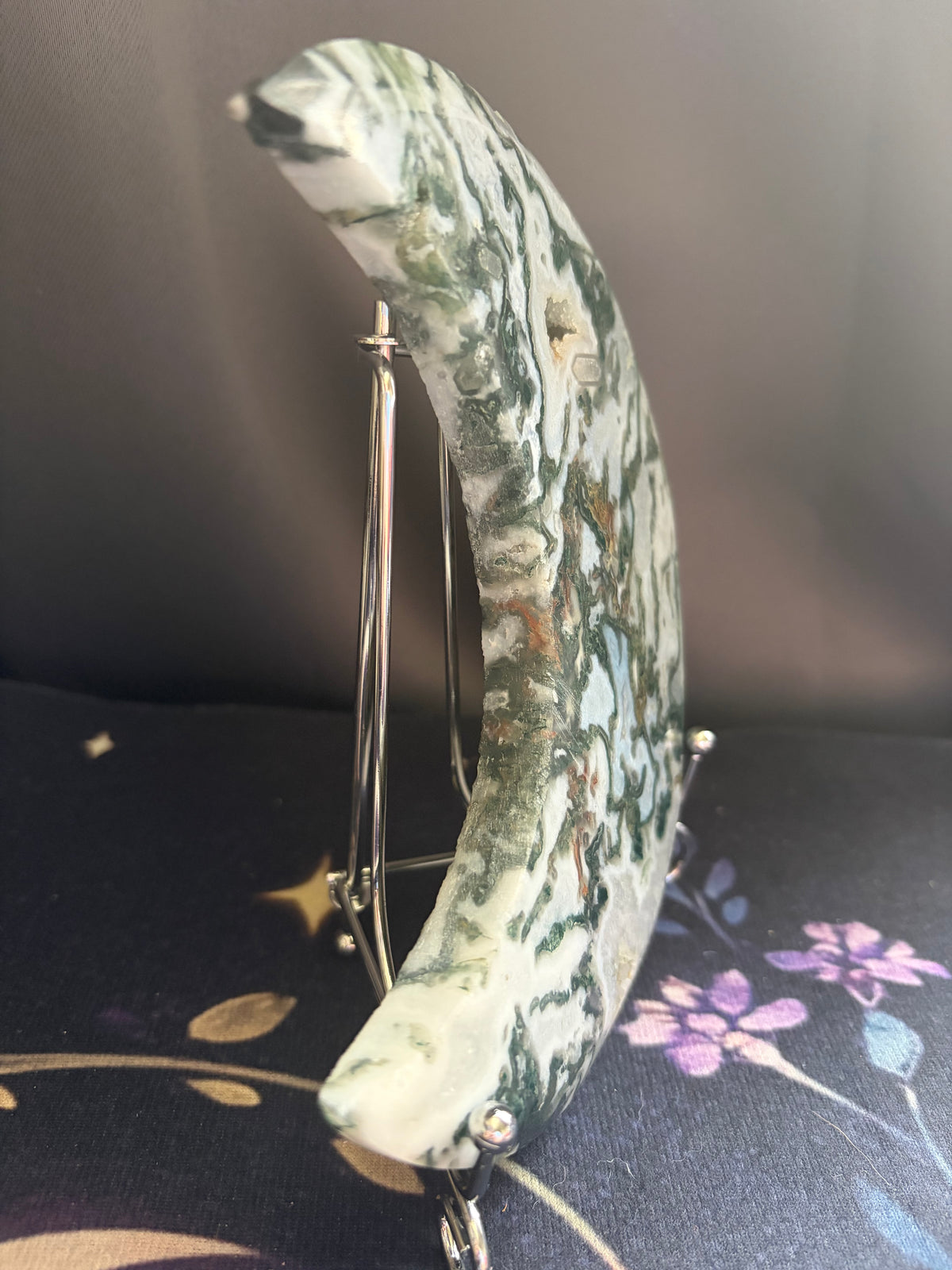 A crescent moon-shaped decorative object made of green and white marbled stone, displayed on a small metal stand against a dark background.