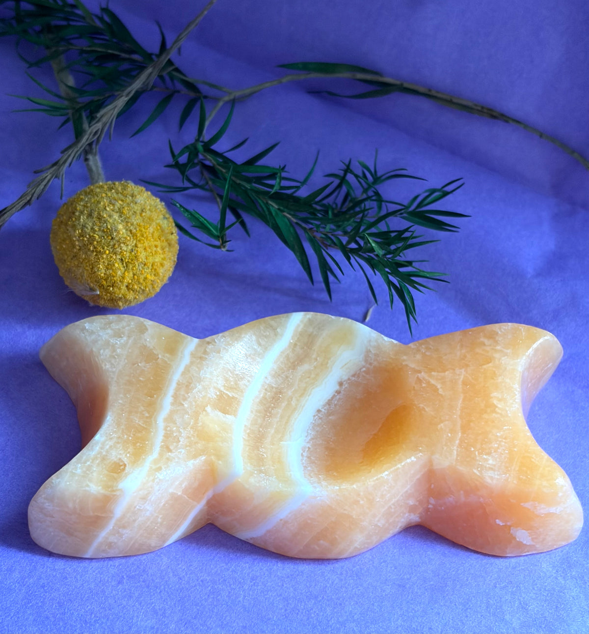 Orange Calcite Triple Moon Goddess Carving displayed on a purple fabric background with green sprigs and a yellow textured ball prop.
