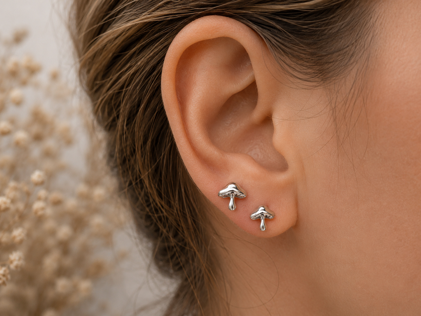 A close-up of a person's ear wearing two small silver mushroom stud earrings, with light brown hair and a soft beige blurred background.