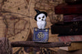 A small decorative owl figurine wearing a black witch hat and perched on a blue book with a gold pentagram, displayed on a rustic wooden branch against a vintage map and stacked books background.