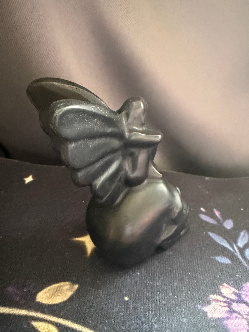 A small black figurine of a winged fairy or angel seated on a dark floral surface, photographed indoors against a soft gray fabric background.