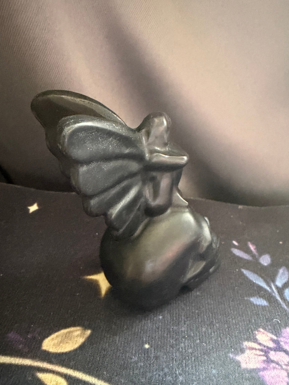 A small black figurine of a winged fairy or angel seated on a dark floral surface, photographed indoors against a soft gray fabric background.
