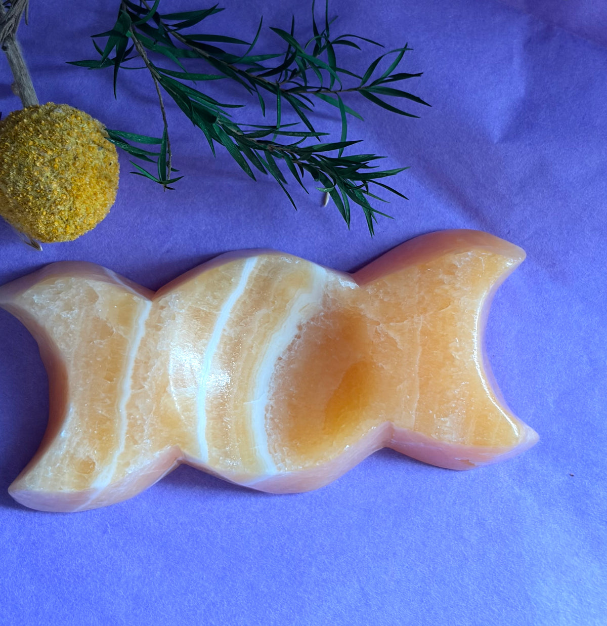 Orange Calcite Triple Moon Goddess Carving displayed on a purple background with decorative greenery and a yellow flower.