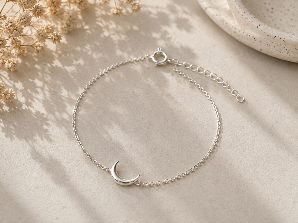 Crescent Moon Chain Bracelet Sterling Silver product shot — delicate silver chain bracelet with small polished crescent moon charm, spring ring clasp and extender chain, on cream stone surface with dried gypsophila"